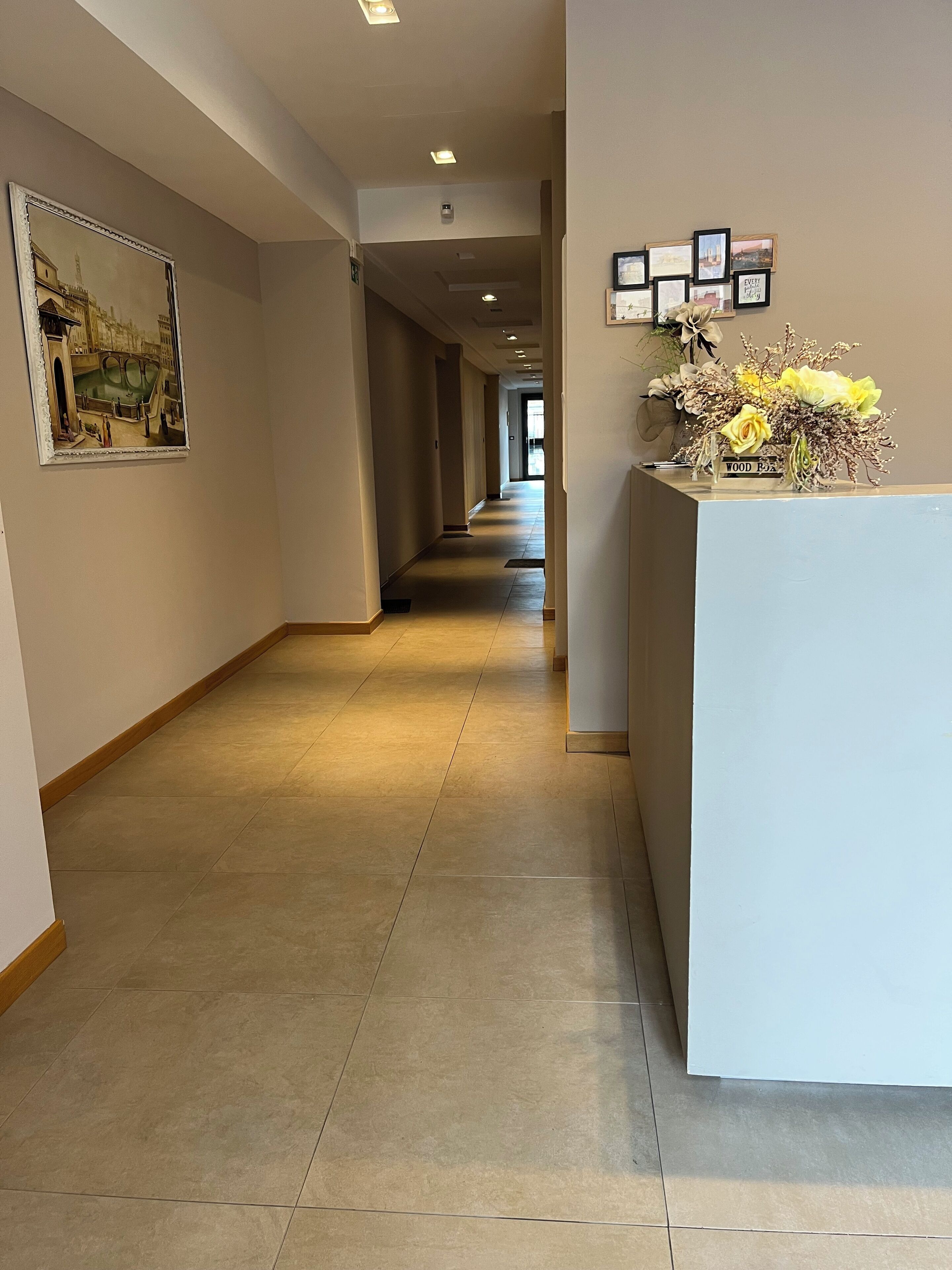 A hotel reception area with a long corridor, a reception desk with a floral arrangement, and framed pictures on the walls.