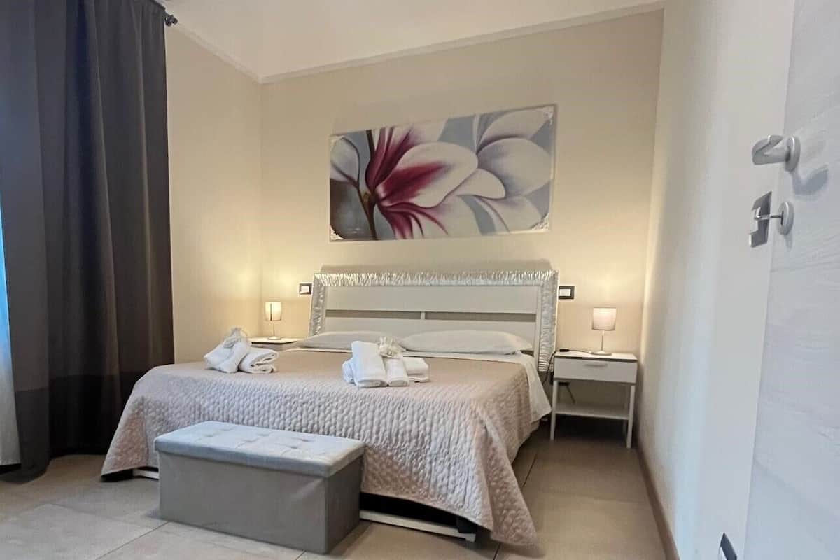 A modern hotel room with a large bed, bedside tables with lamps, a bench, and a floral artwork on the wall.