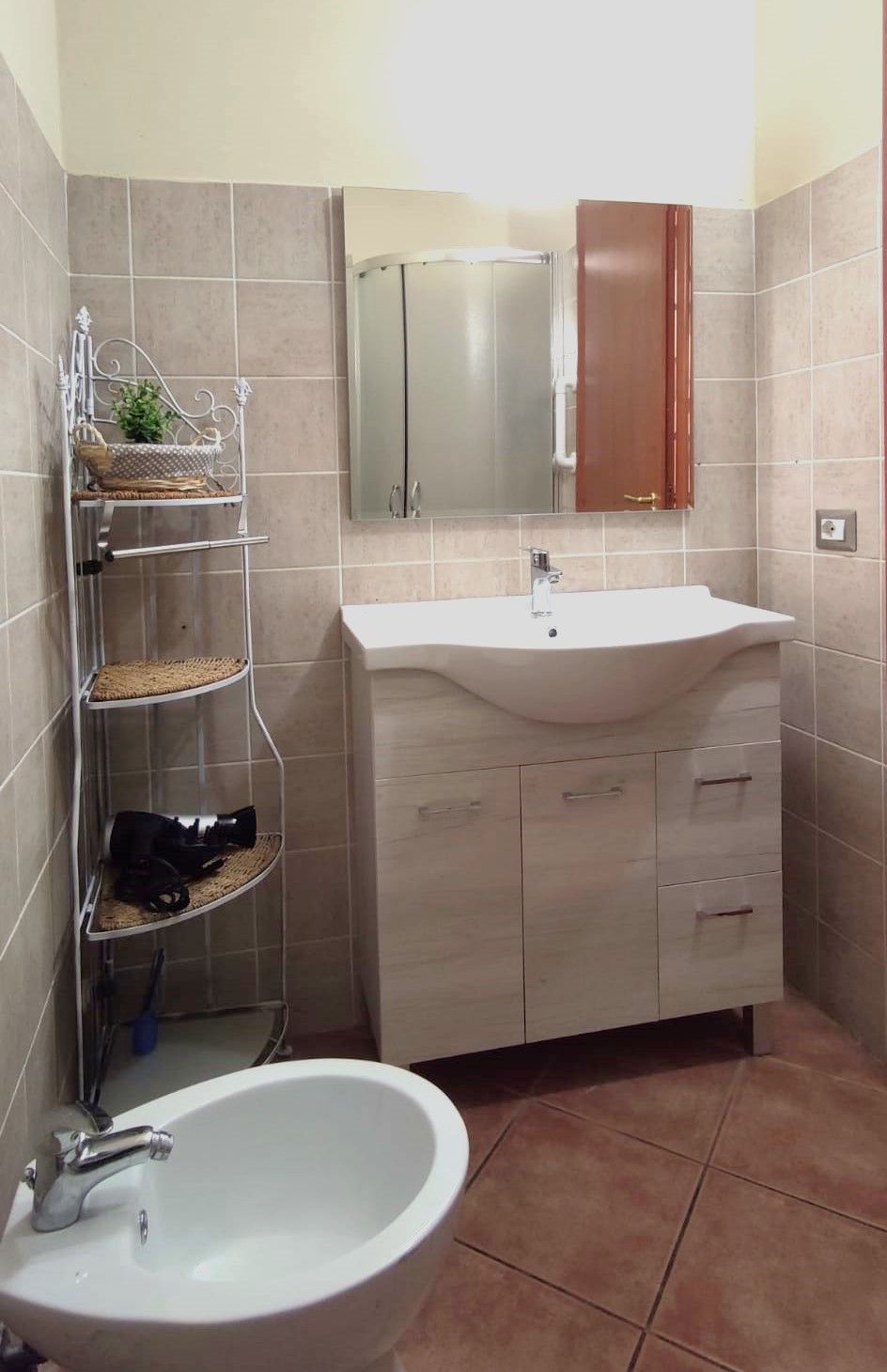 Une salle de bain avec un lavabo blanc, un miroir au-dessus et un meuble en dessous. On y trouve une étagère avec des paniers et une paire de chaussures.
