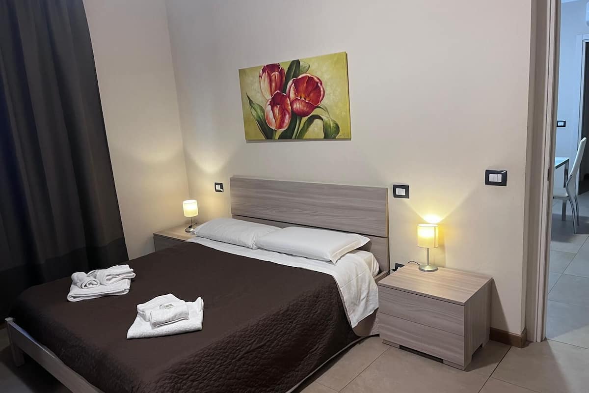 A hotel room with a large bed, two bedside tables with lamps, a painting of flowers on the wall, and a view of the hallway.
