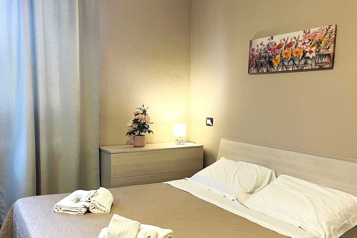 A hotel room with a bed, a nightstand, a lamp, a painting, and a window with curtains.