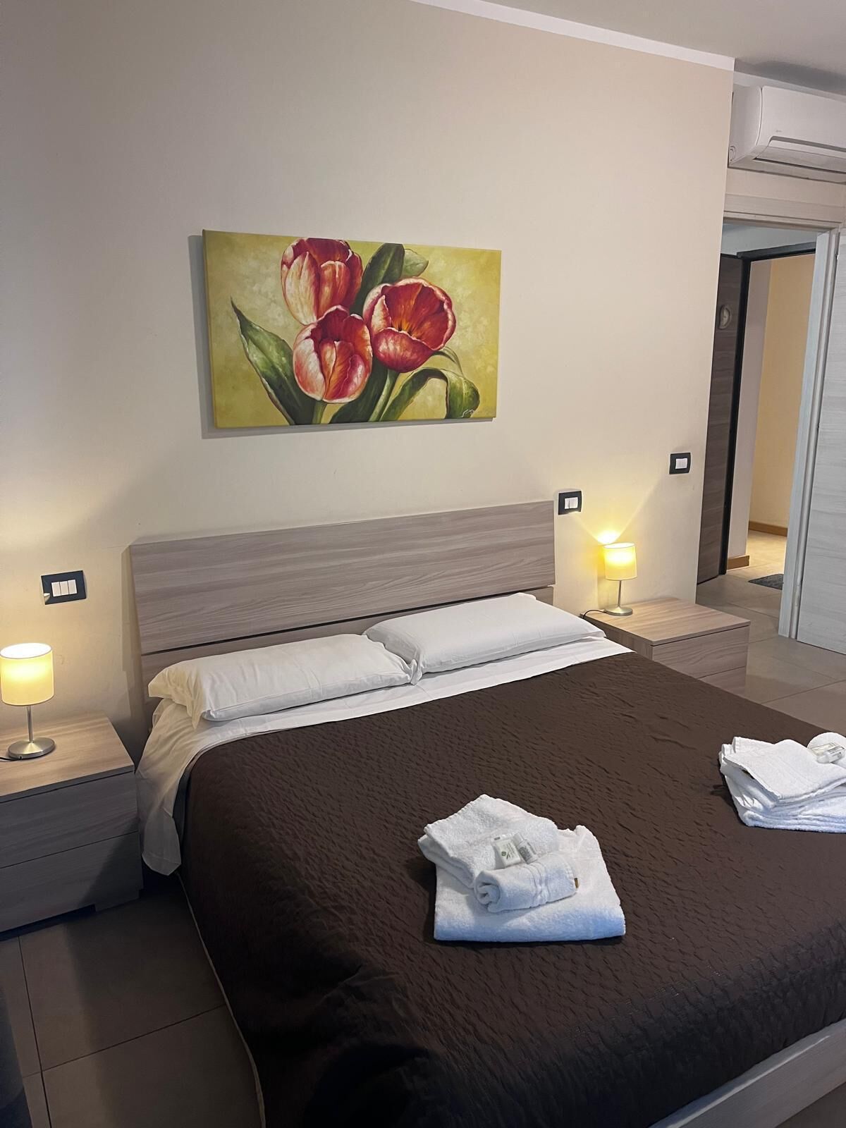 A neatly made bed with white linens, two bedside lamps, and a painting of red tulips on the wall.