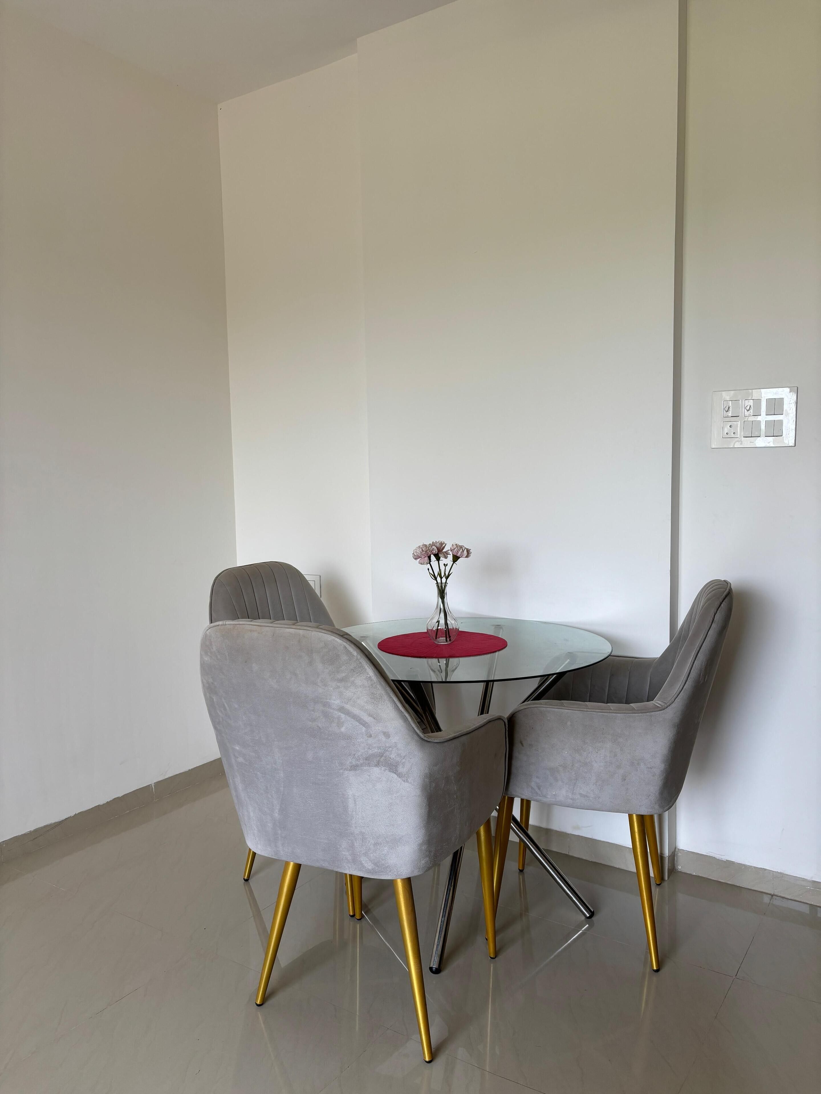 Zona pranzo con tavolo in vetro, due poltrone grigie con gambe dorate e un vaso con fiori sul tavolo.