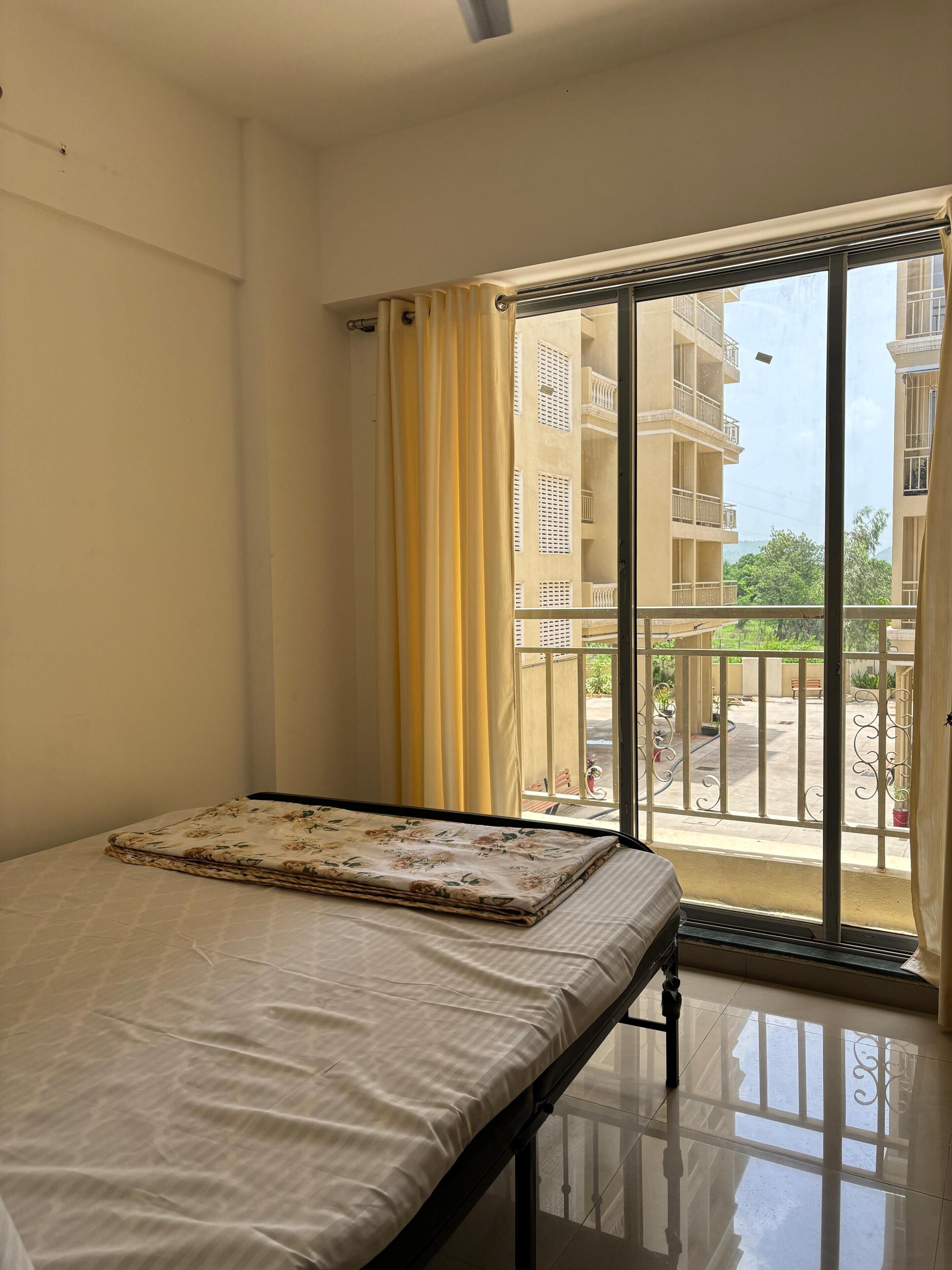 A room with a bed, a window with a view of buildings and greenery, and a balcony with a railing.