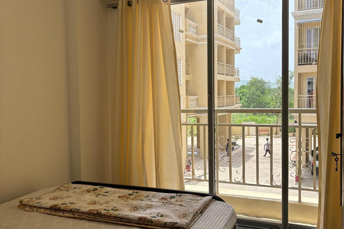 Quarto com cama, janela com cortinas e vista para uma varanda e edifícios.