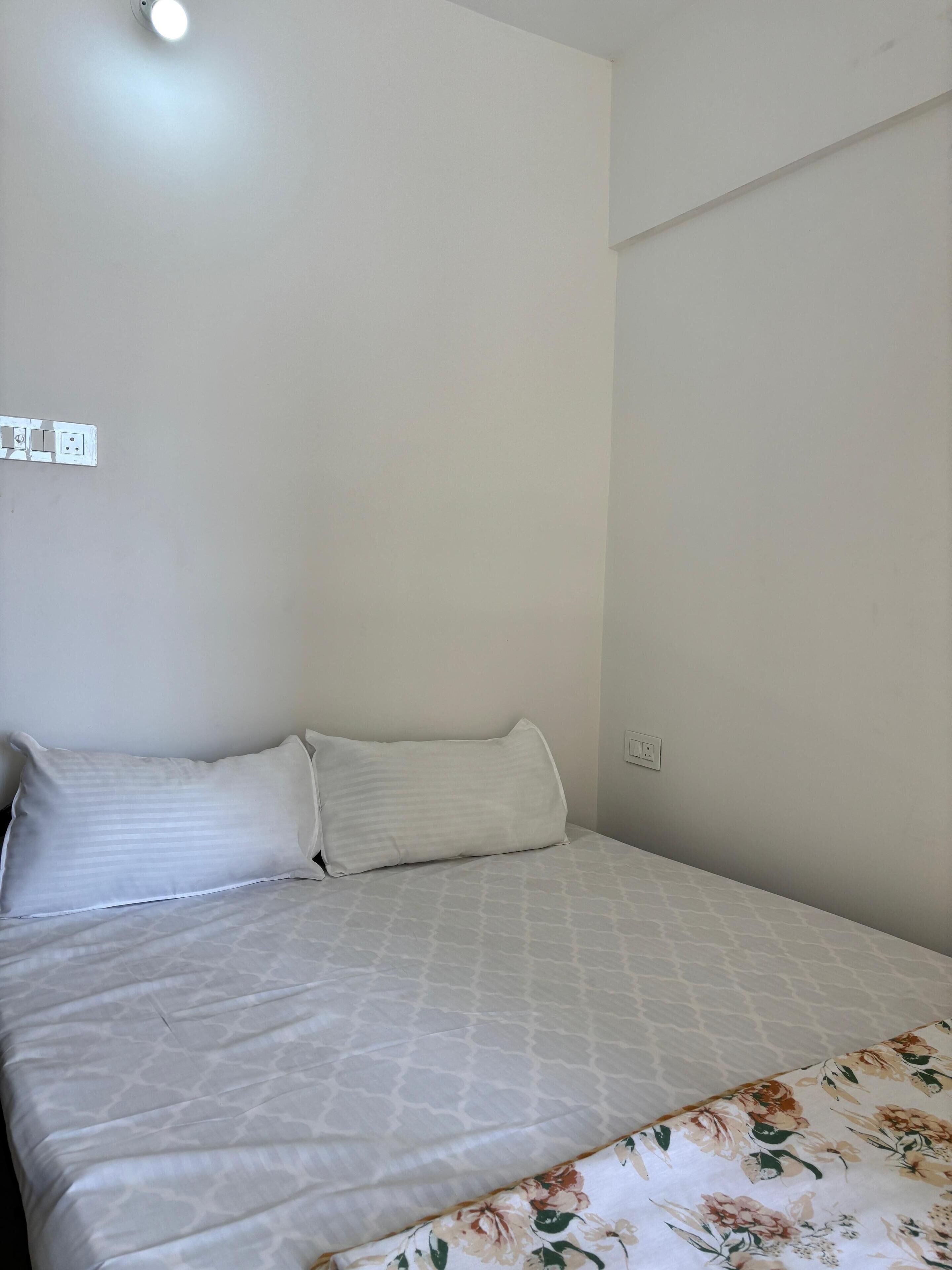 A bed with a patterned bedspread and two pillows, a wall socket, and a ceiling light fixture.