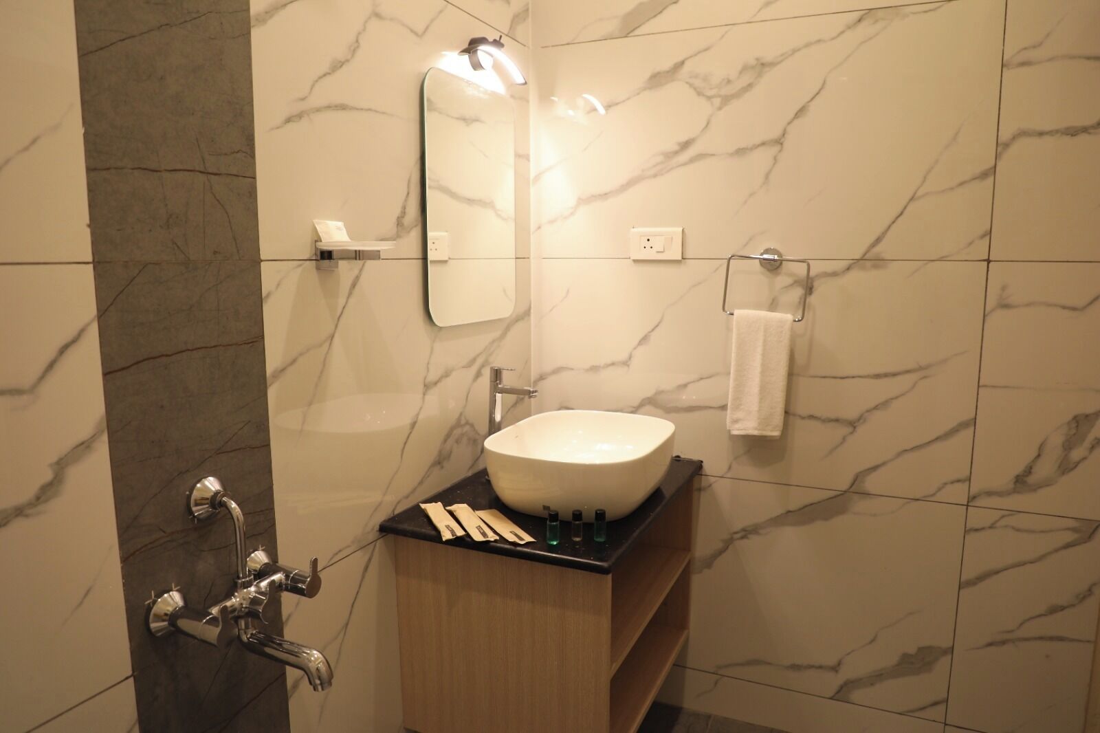 A bathroom with a marble wall, a sink with a mirror above it, a towel hanging on a rack, and a shower area.