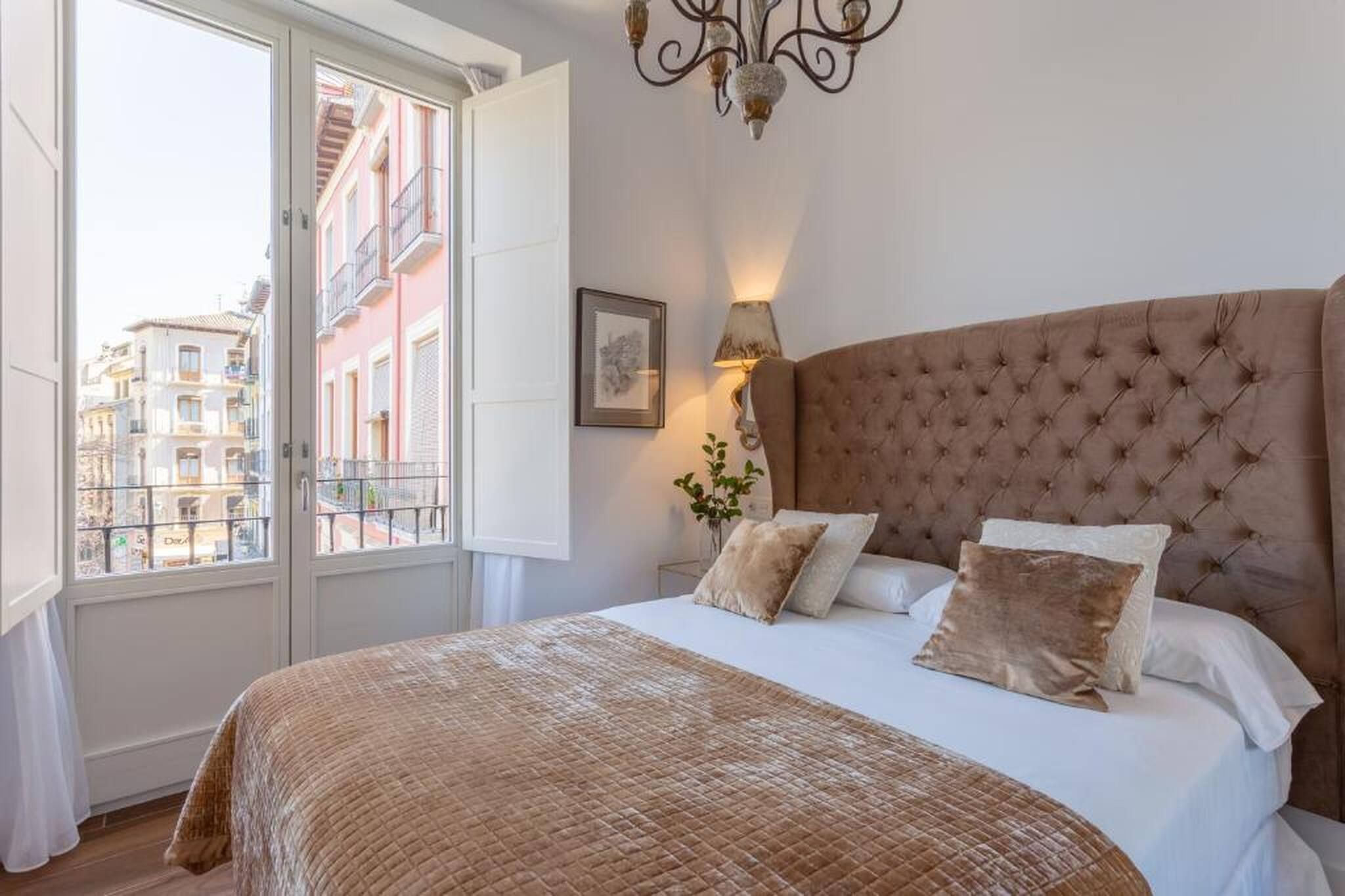 A bedroom with a large bed, a window with a view of buildings, a wall-mounted lamp, and a framed picture.