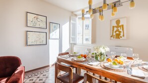A dining area with a wooden table set for four, surrounded by chairs. There are framed artworks on the wall, a radiator, and a window with curtains. - Casa Dante in Lucca With 3 Bedrooms and 2 Bathrooms (Lucca)