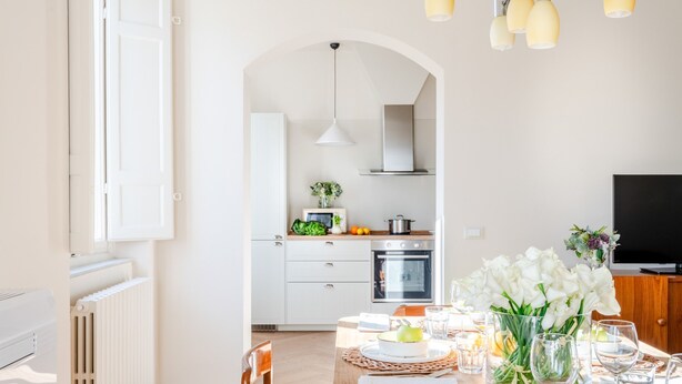 A modern kitchen with white cabinets, a stove, and an oven. There is a dining table with a vase of flowers and a bowl of fruit. - Casa Dante in Lucca With 3 Bedrooms and 2 Bathrooms (Lucca)