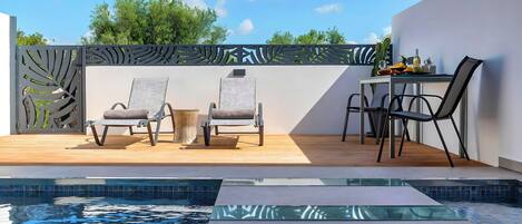 A modern outdoor pool area with a wooden deck, two lounge chairs, a table, and a glass-topped table.