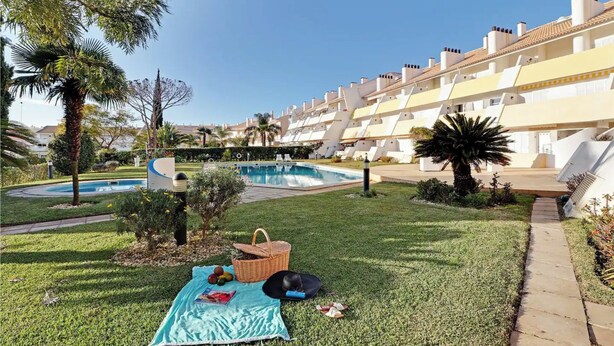 A poolside area with a grassy lawn, a picnic setup, and a row of white buildings with balconies. - Marina Family Duplex with Pool (Loulé)