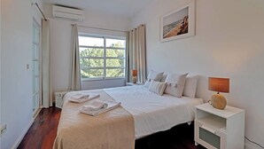 A bedroom with a large bed, a bedside table with a lamp, a window with curtains, and a painting on the wall. - Marina Family Duplex with Pool (Loulé)