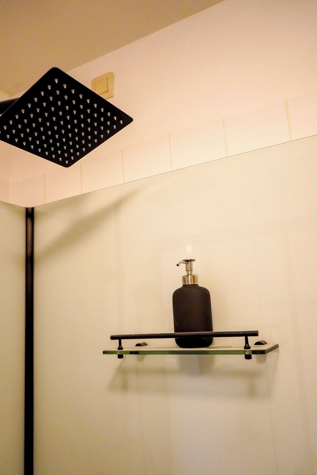Een badkamer met een zwarte douchekop, een zwarte fles op een glazen plank en een witte betegelde muur.