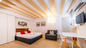 A modern hotel room with a bed, a sofa, a dining table, and a TV.