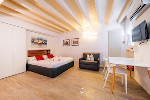 A modern hotel room with a bed, a sofa, a dining table, and a TV.