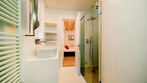 A modern bathroom with a glass shower enclosure, a white sink, and a small bedside table with a red pillow.