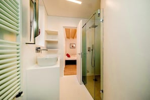 A modern bathroom with a glass shower enclosure, a white sink, and a small bedside table with a red pillow.