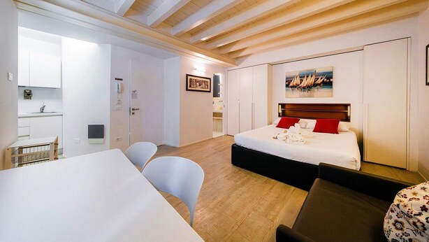 A hotel room with a bed, a desk, chairs, a small kitchenette, and a painting on the wall.