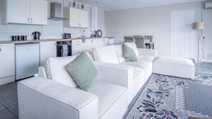 Una sala de estar moderna con un conjunto de sofá, un comedor y una cocina con armarios blancos y horno empotrado.