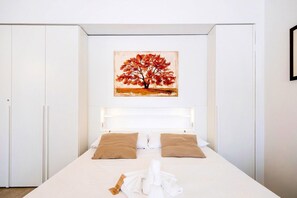 A bedroom with a bed, white headboard, two pillows, a painting of a tree, and a towel on the bed.