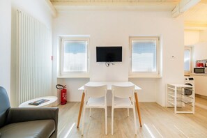 A modern living room with a flat-screen TV, a dining table with chairs, a sofa, a fire extinguisher, and a kitchen area with a microwave.