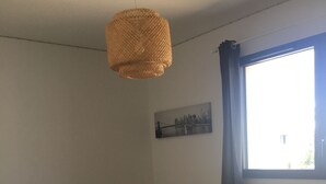 Ett litet, välstädat rum med en säng, ett litet bord, en hängande lampa och ett fönster med gardiner.