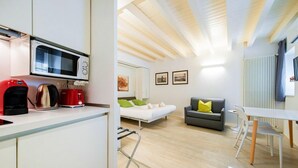 A modern kitchen with a microwave, coffee maker, and kettle. There is a bed with green pillows, a gray sofa with a yellow pillow, and a dining area with white chairs. - ApartmentsArena - Zini Studios - 1 (Verona)