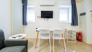 A compact room with a white table, two white chairs, a grey sofa, a mounted TV, a radiator, a window with curtains, and a fire extinguisher. - ApartmentsArena - Zini Studios - 1 (Verona)