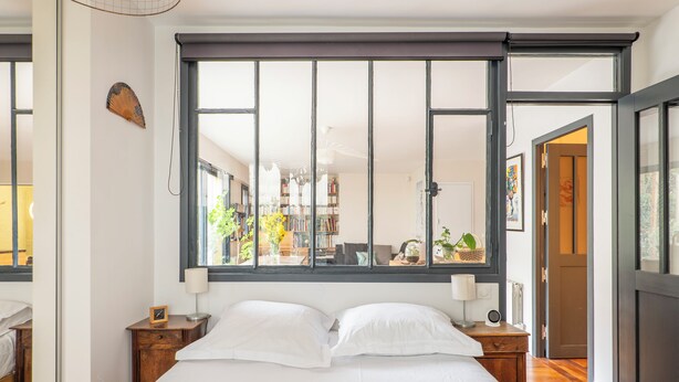 A bedroom with a large window, a bed with white linens, wooden nightstands, and a view of a library and garden.