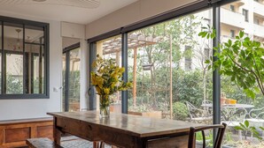 A wooden table with benches, large windows, and a view of greenery outside.
