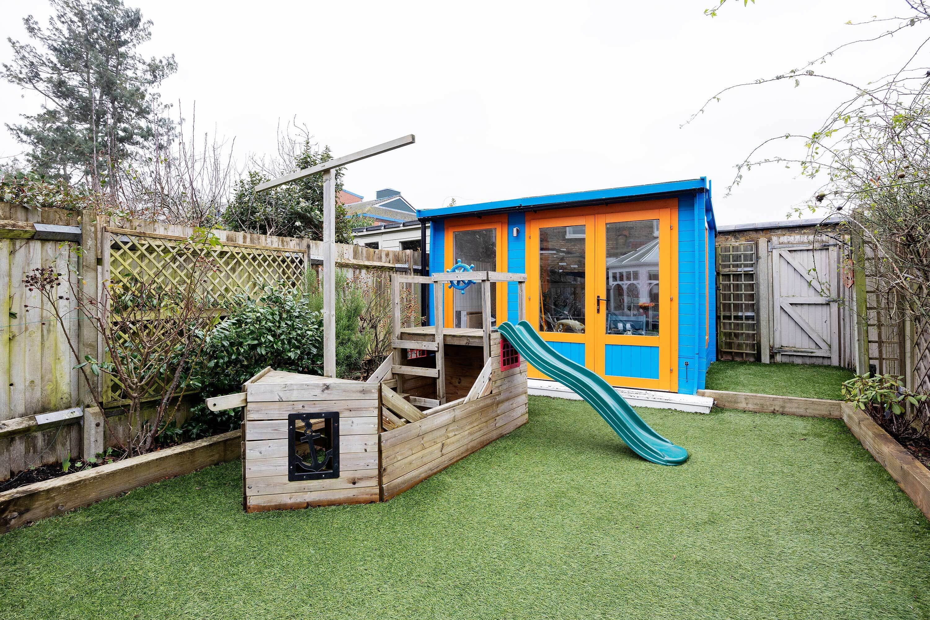 Una casita de juegos de madera con un tobogán, una pequeña terraza y un jardín vallado.