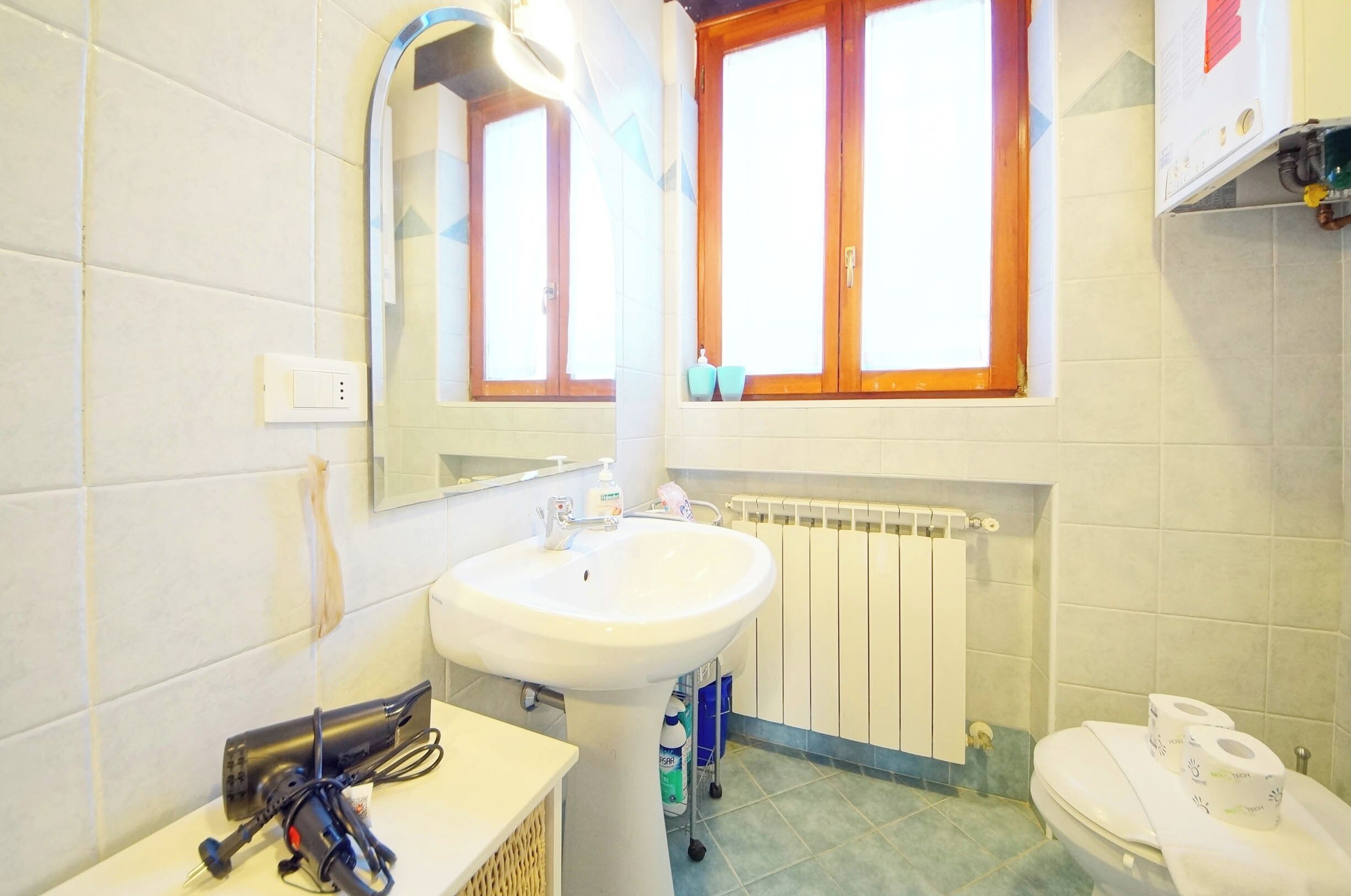 A bathroom with a white sink, a mirror, a window with wooden frames, a toilet, and a radiator.