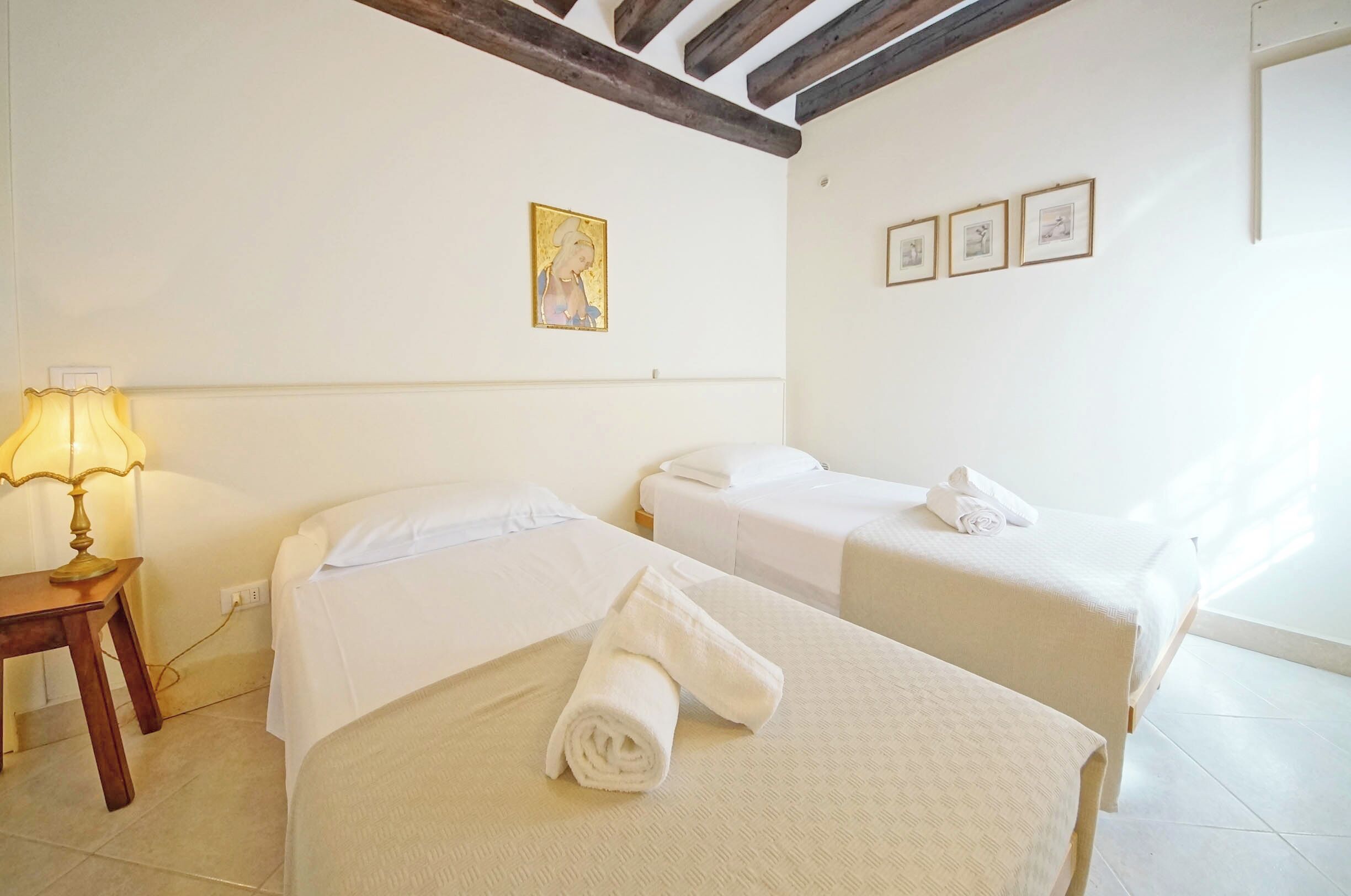 A hotel room with two beds, a bedside table with a lamp, and a wall with framed pictures.