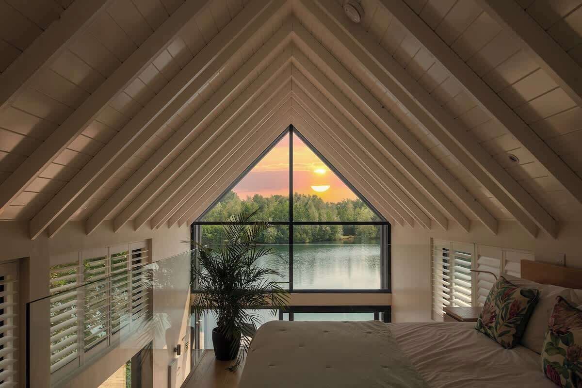 Un dormitorio con una ventana grande, una cama, una planta en maceta y vistas a un lago al atardecer.