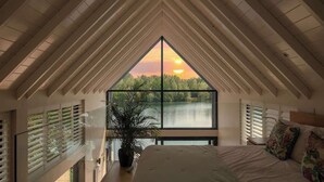 A bedroom with a large window, a bed, a potted plant, and a view of a lake at sunset. - The Boathouse - Romantic Glass Lodge - Hot Tub (South Cerney)
