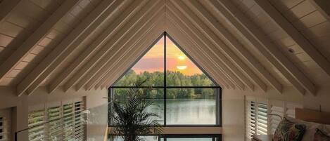 Un dormitorio con una ventana grande, una cama, una planta en maceta y vistas a un lago al atardecer.