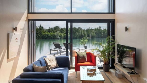 A modern living room with a large window overlooking a lake, a sofa, a red armchair, a coffee table, and a flat-screen TV. - The Boathouse - Romantic Glass Lodge - Hot Tub (South Cerney)