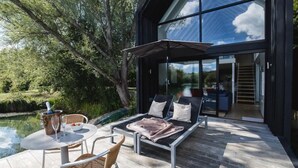A modern outdoor seating area with a glass enclosure, a table with chairs, and a view of the surrounding greenery. - The Boathouse - Romantic Glass Lodge - Hot Tub (South Cerney)