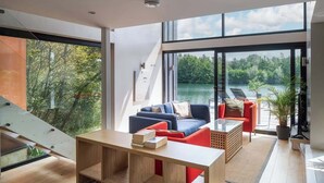 A modern living room with a large window, a staircase, and a view of a lake. - The Boathouse - Romantic Glass Lodge - Hot Tub (South Cerney)