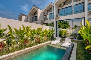 A modern building with a swimming pool, surrounded by lush greenery and a clear blue sky.