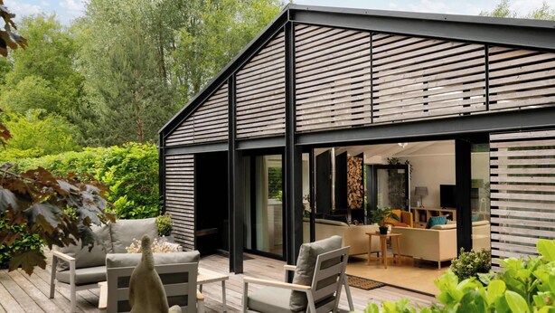 Ein modernes Haus mit einer Terrasse, Sitzgelegenheiten im Freien und einem einsehbaren Innenraum mit Sofa und Essbereich.