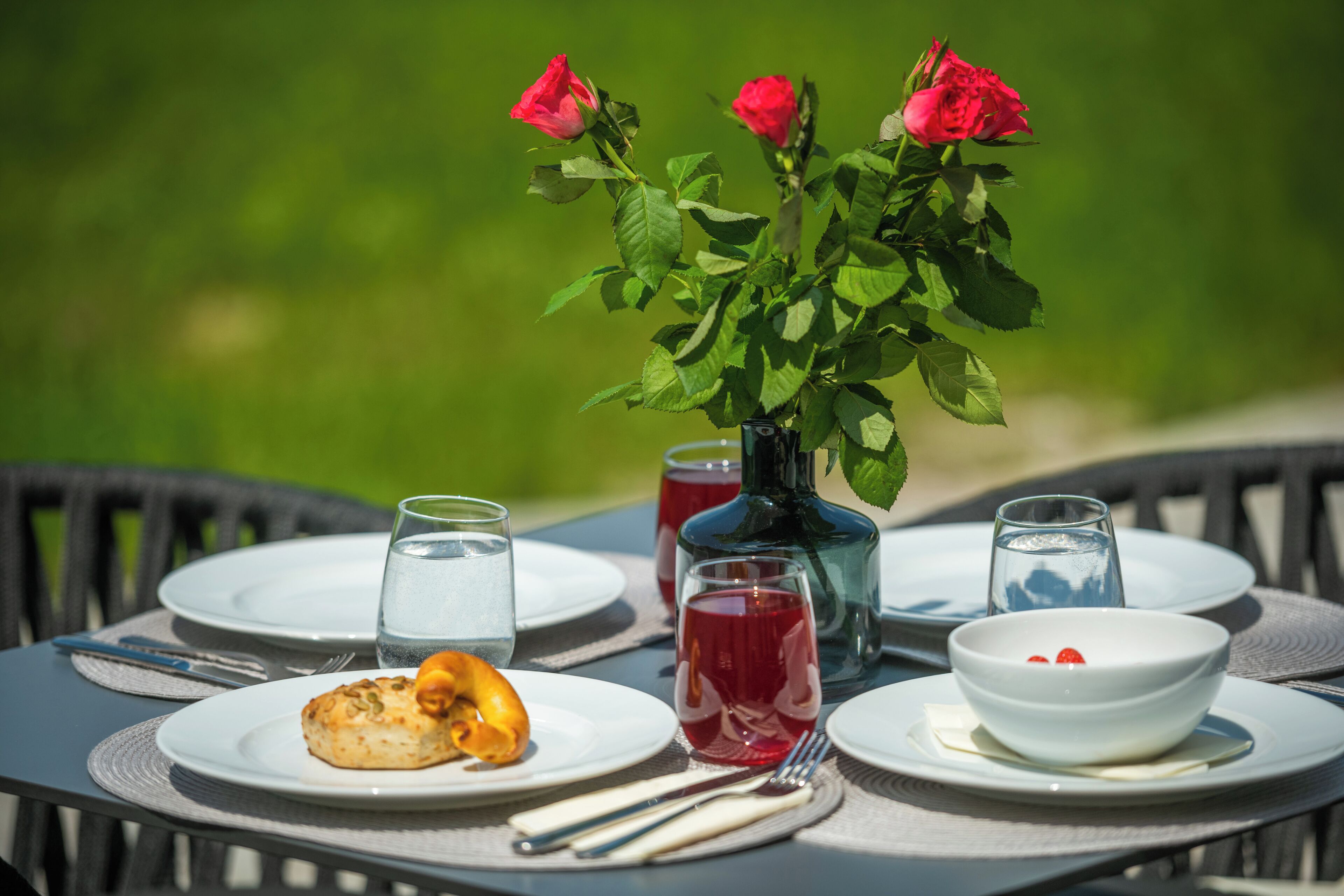 Una mesa lista para comer, con platos, cubiertos, vasos y un jarrón con rosas rojas.