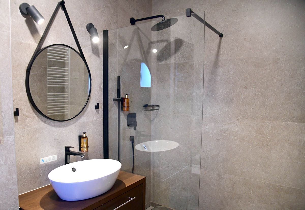 Un baño moderno con un espejo redondo, un lavamanos blanco y una zona de ducha con mampara de vidrio.
