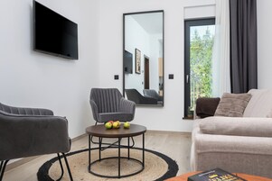 Un moderno salón con un televisor de pantalla plana, un espejo grande, un sillón gris, una mesa de centro redonda con frutas y un sofá beige.
