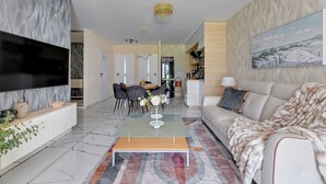 En moderne stue med et fladskærms-tv, et sofabord i glas, en sofa og et spiseområde med stole og et bord.