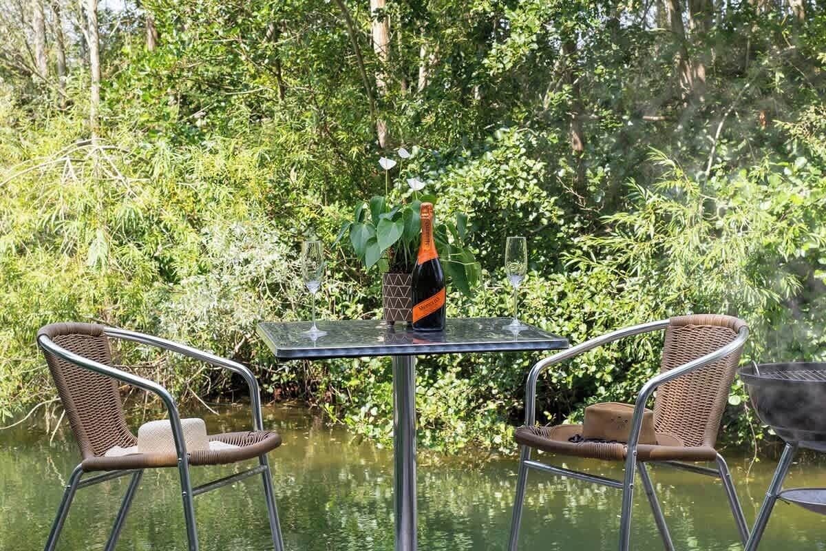Una mesa con una botella y vasos, rodeada de sillas de mimbre, cerca de un cuerpo de agua con vegetación al fondo.