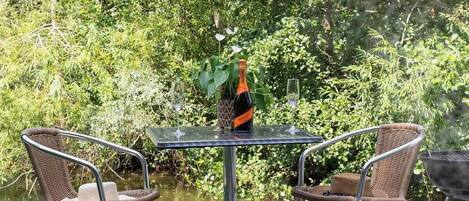 Una mesa con una botella y vasos, rodeada de sillas de mimbre, cerca de un cuerpo de agua con vegetación al fondo.
