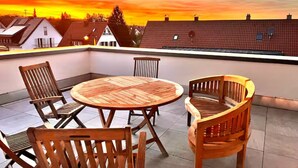 A rooftop terrace with a round wooden table and four wooden chairs, overlooking a residential area during sunset.