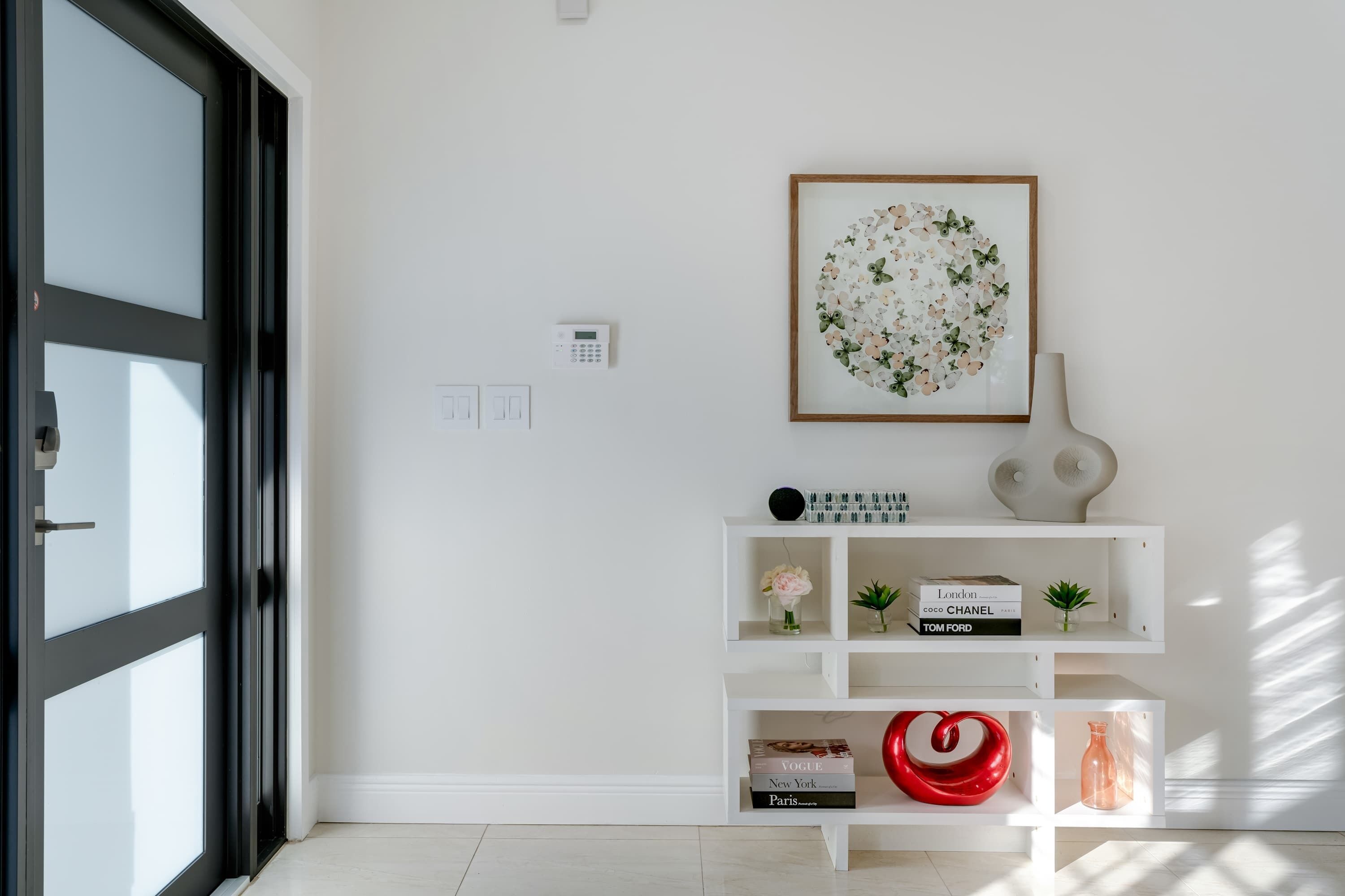 A modern interior with a white wall, a framed floral artwork, a white console table with books and decorative items, and a black-framed door.